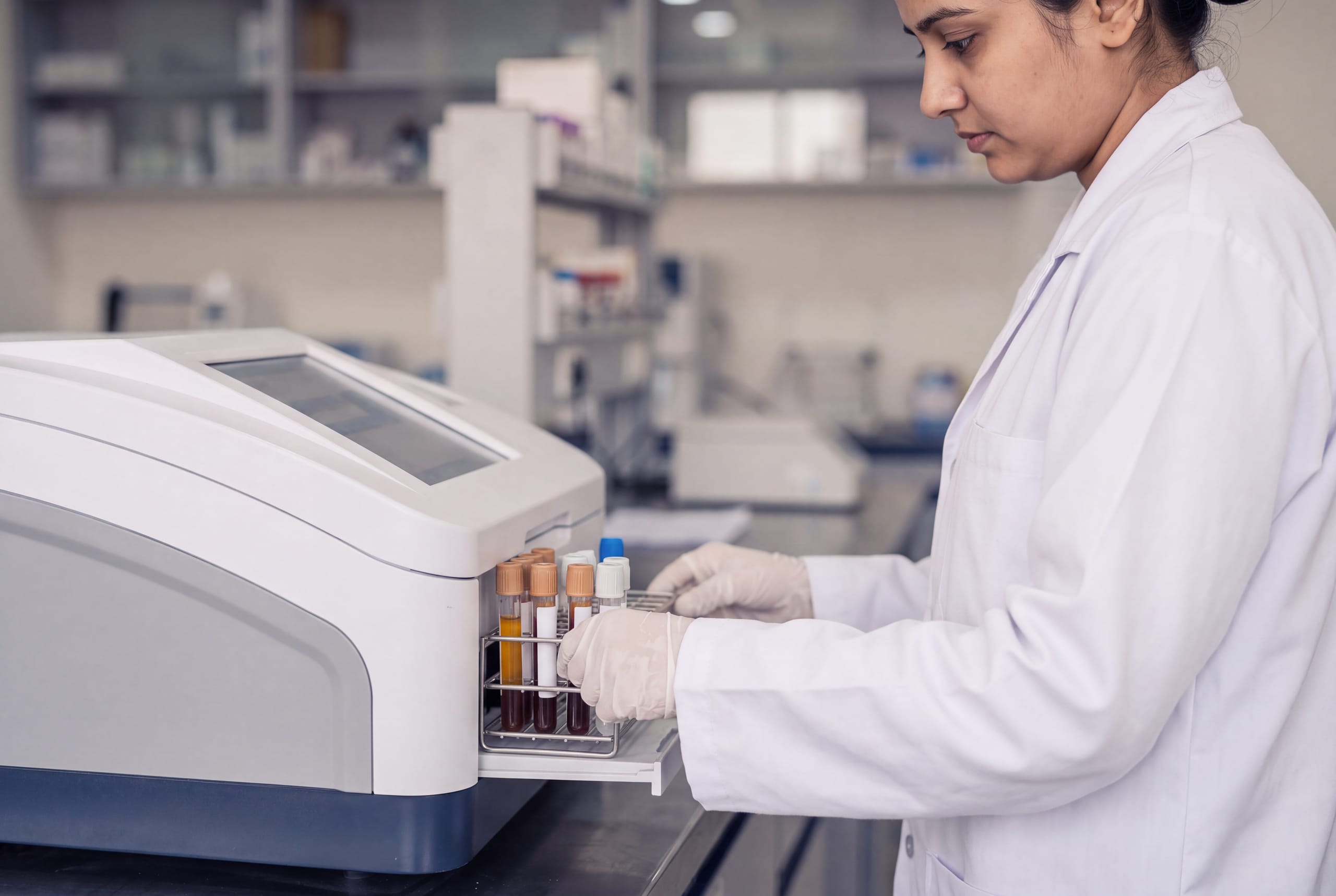 A technologist in a white coat loading a rack of capped tubes into a benchtop analyzer.